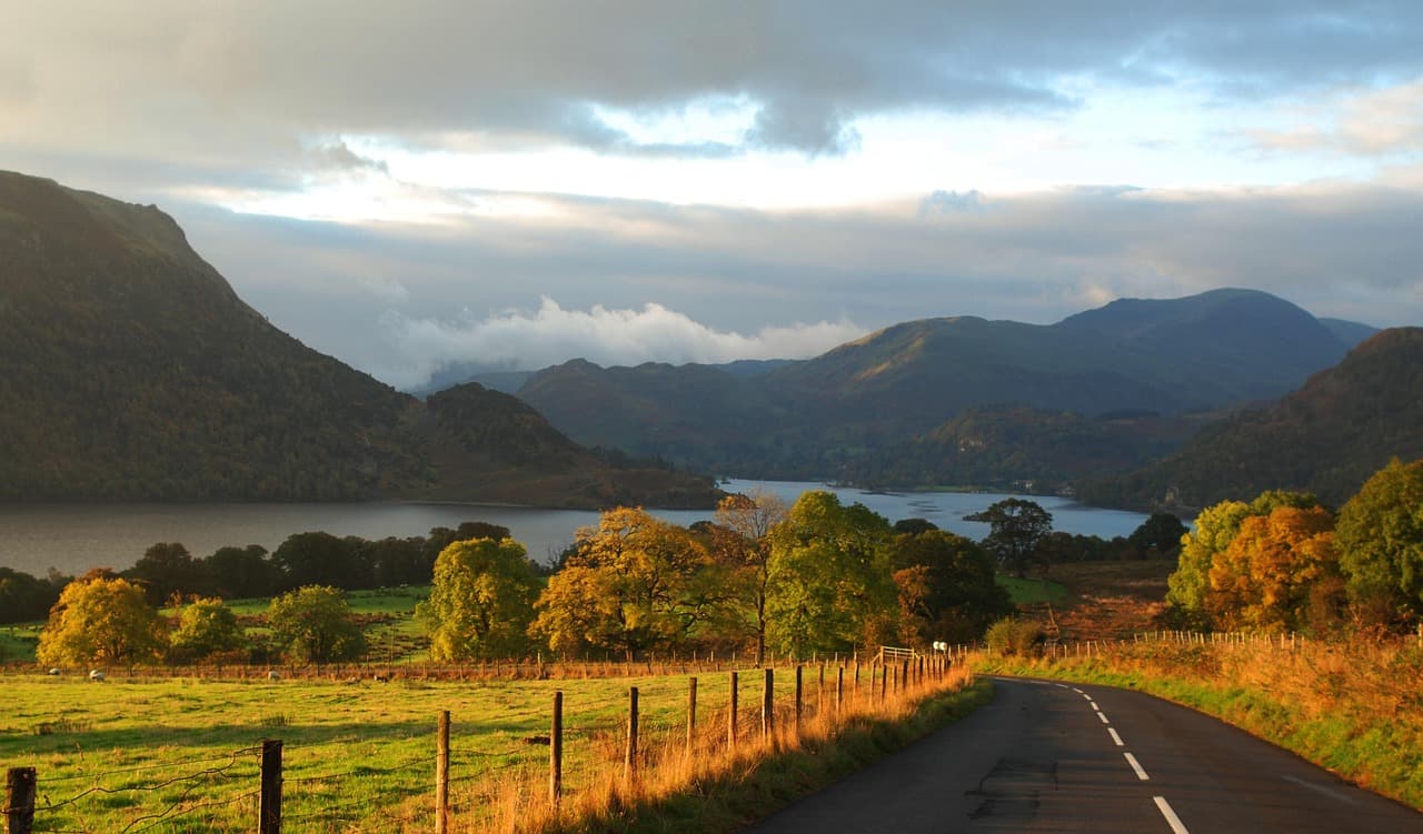 A country road curving past autumn trees and a lake beneath the fells